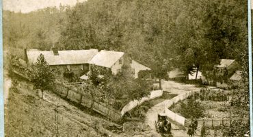Sepia toned image of a mountain inn in the late 19th century. Travelers with a carriage are shown in the foreground.