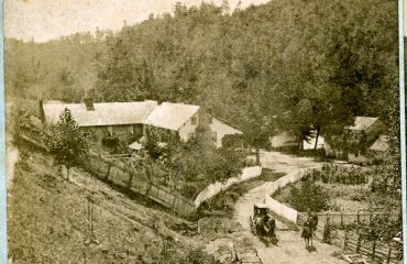 Sepia toned image of a mountain inn in the late 19th century. Travelers with a carriage are shown in the foreground.